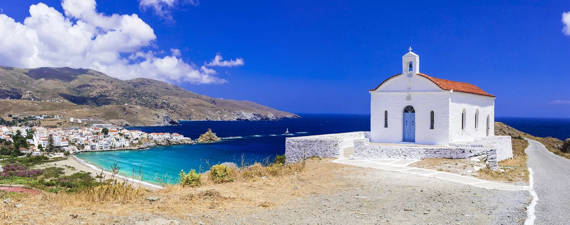 Chapelle blanche dominant une baie turquoise et un village côtier - Andros - Cyclades